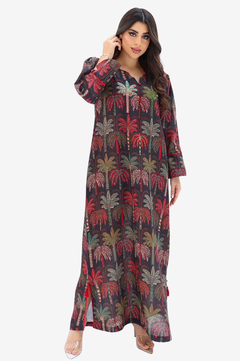 A woman modeling a long-sleeved maxi dress with a dark charcoal base and an all-over palm tree print in red, green, and beige, set against a plain light gray background.