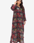 A woman modeling a long-sleeved maxi dress with a dark charcoal base and an all-over palm tree print in red, green, and beige, set against a plain light gray background.