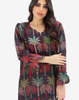 A woman modeling a long-sleeve tunic dress with a dark background and a colorful palm tree print in red, green, and beige, set against a plain light gray background.