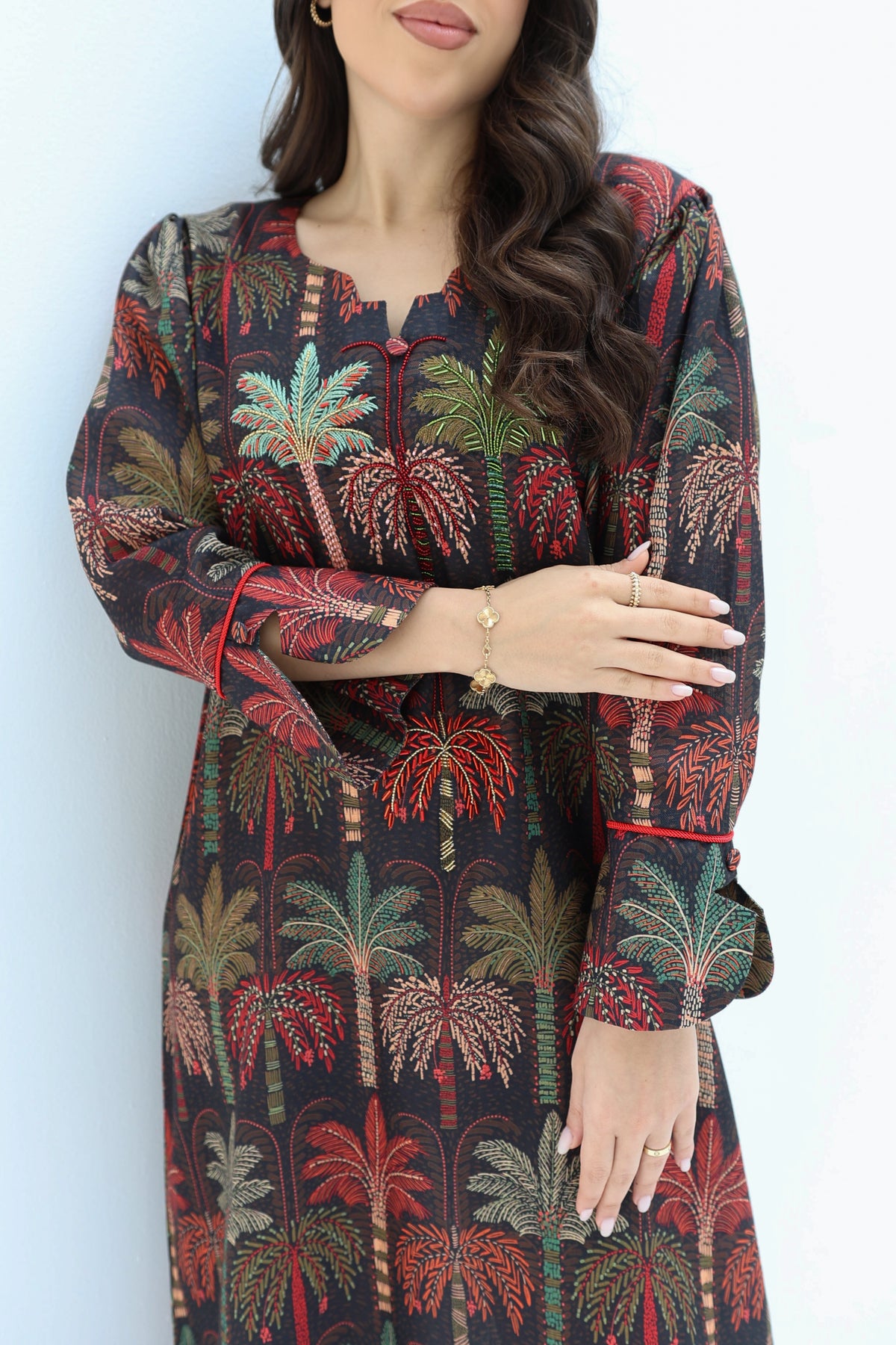 A woman wearing a long-sleeve tunic dress with a tropical palm tree print on a dark background. The dress features red, green, and beige palm trees with red piping details along the collar, cuffs, and front button placket.