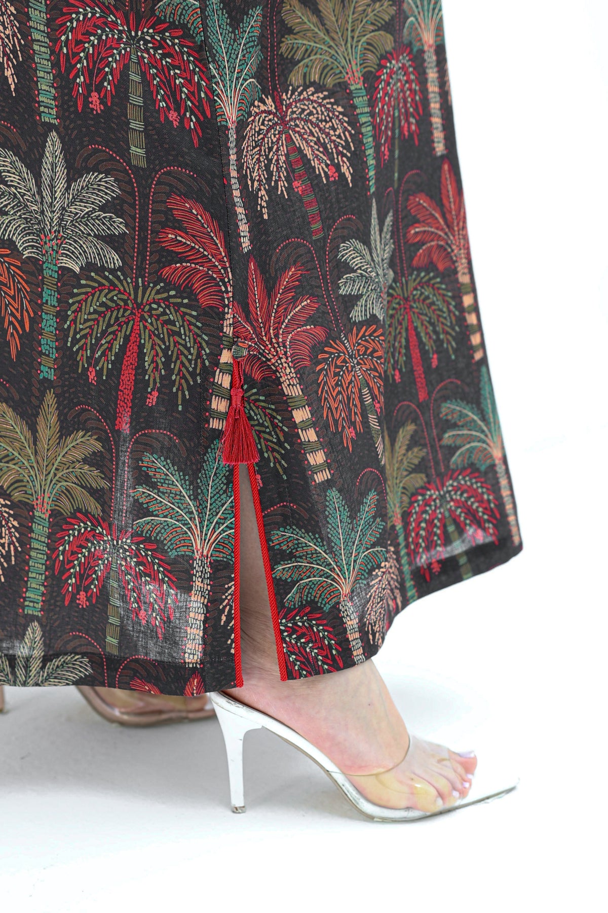 Close-up of a person wearing a black maxi skirt with a red, green, and beige palm tree print, paired with clear high-heeled sandals against a white background.
