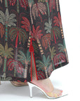 Close-up of a person wearing a black maxi skirt with a red, green, and beige palm tree print, paired with clear high-heeled sandals against a white background.