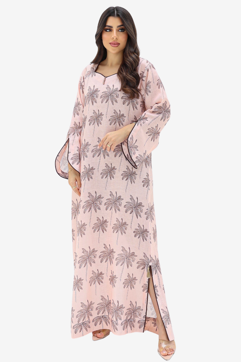 A woman modeling a long light pink kaftan dress with a gray palm tree pattern, shown against a plain light gray background.