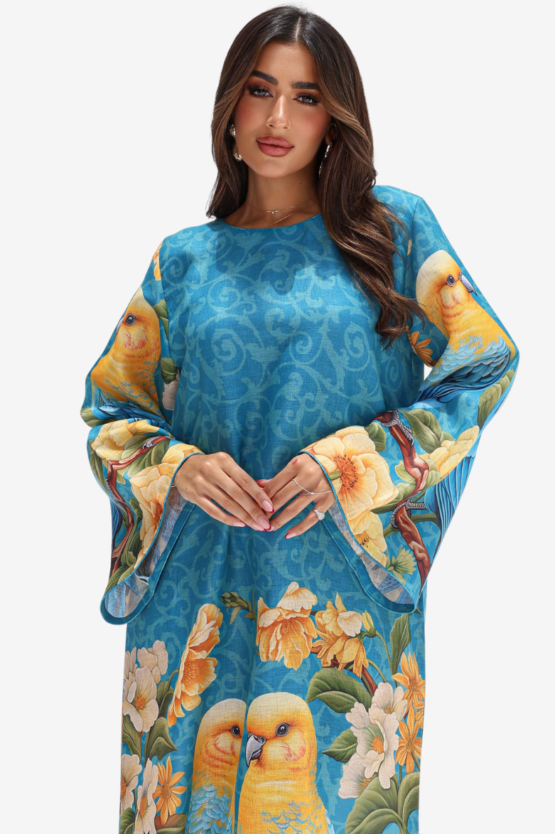 A woman modeling a vibrant blue kaftan dress with a printed pattern of yellow birds and large yellow and white flowers, set against a plain light gray background.