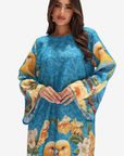 A woman modeling a vibrant blue kaftan dress with a printed pattern of yellow birds and large yellow and white flowers, set against a plain light gray background.