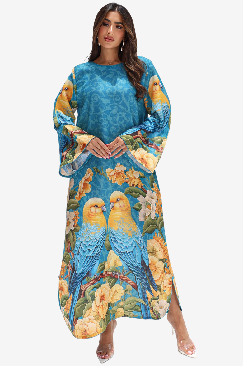 A woman modeling a long-sleeved maxi dress with a vibrant blue base and a detailed print of blue and yellow parrots, green leaves, and orange flowers, set against a plain light gray background.