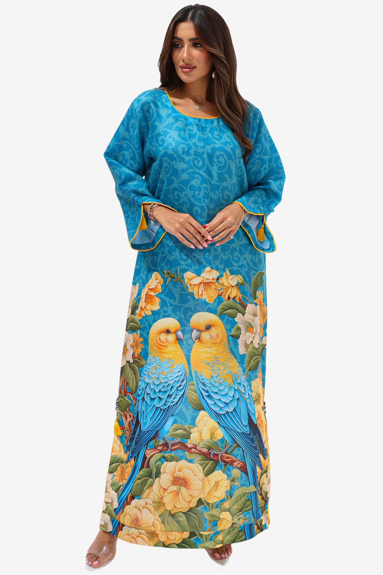 A woman modeling a long blue kaftan dress featuring a yellow and blue parrot and yellow floral print, set against a plain light gray background.