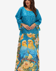 A woman modeling a long blue kaftan dress featuring a yellow and blue parrot and yellow floral print, set against a plain light gray background.