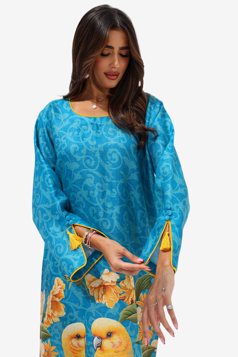 A woman wearing a bright blue tunic dress with a subtle tonal swirl pattern, yellow floral details, and two yellow birds near the hem, shown against a plain light gray background.