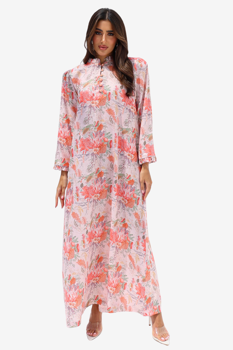 A woman modeling a long-sleeved, high-neck maxi dress with a pink, coral, and green floral print on a light background, set against a plain light gray studio background.