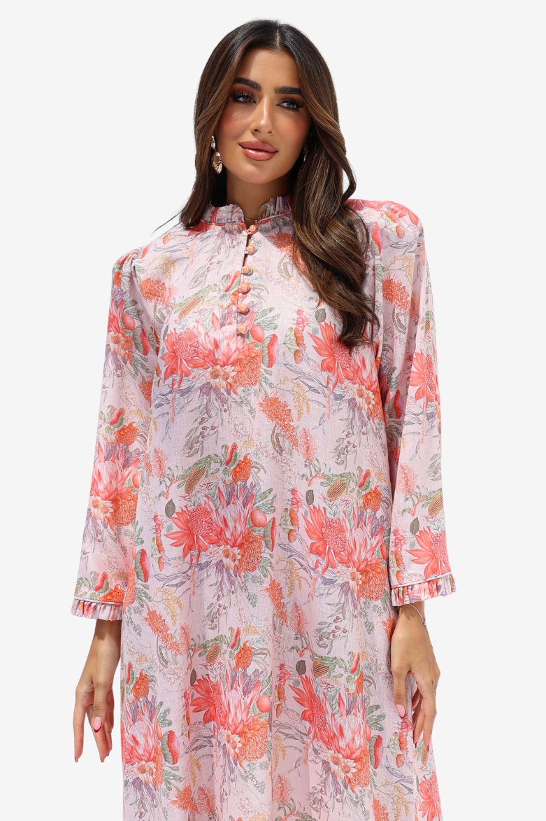 A woman modeling a long-sleeved, high-neck tunic dress with a pink and coral floral print on a light background, set against a plain light gray studio backdrop.