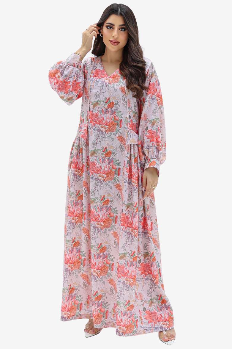 A woman modeling a long-sleeved, maxi-length floral dress with a pink and coral print on a light lavender background, set against a plain light gray studio backdrop.