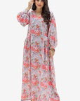 A woman modeling a long-sleeved, maxi-length floral dress with a pink and coral print on a light lavender background, set against a plain light gray studio backdrop.