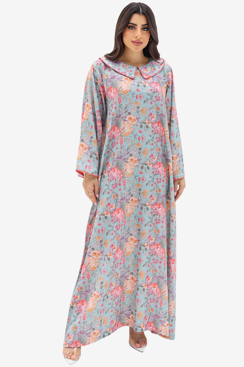 A woman modeling a long, loose-fitting maxi dress with a light blue-gray base and an all-over pink, peach, and beige floral print, set against a plain light gray background.