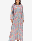 A woman modeling a long, loose-fitting maxi dress with a light blue-gray base and an all-over pink, peach, and beige floral print, set against a plain light gray background.