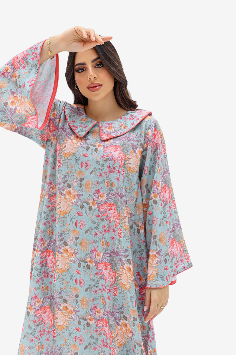 A woman wearing a long-sleeved kaftan dress with a light blue-gray base and a colorful floral print in pink, orange, and purple, featuring a large Peter Pan collar and red piping details.