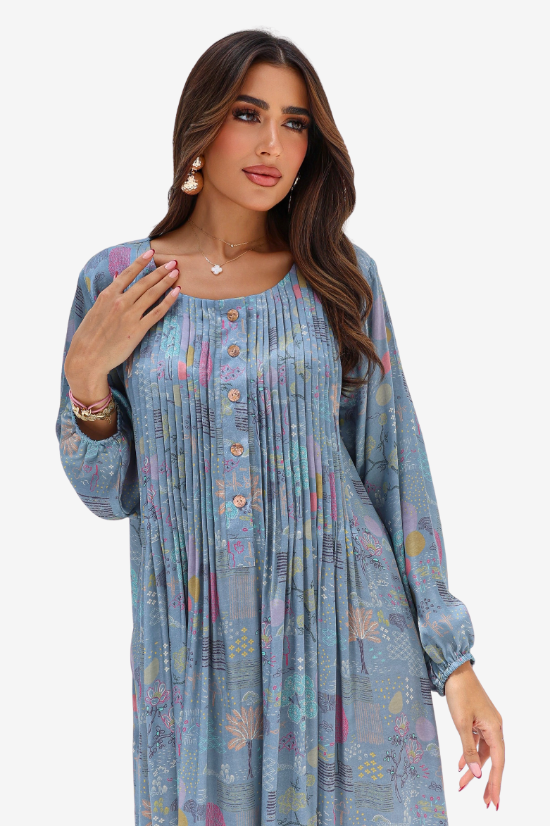 A woman modeling a long-sleeve, pleated tunic dress in a dusty blue color with a subtle multicolor floral and geometric print, set against a plain light gray background.