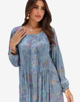 A woman modeling a long-sleeve, pleated tunic dress in a dusty blue color with a subtle multicolor floral and geometric print, set against a plain light gray background.