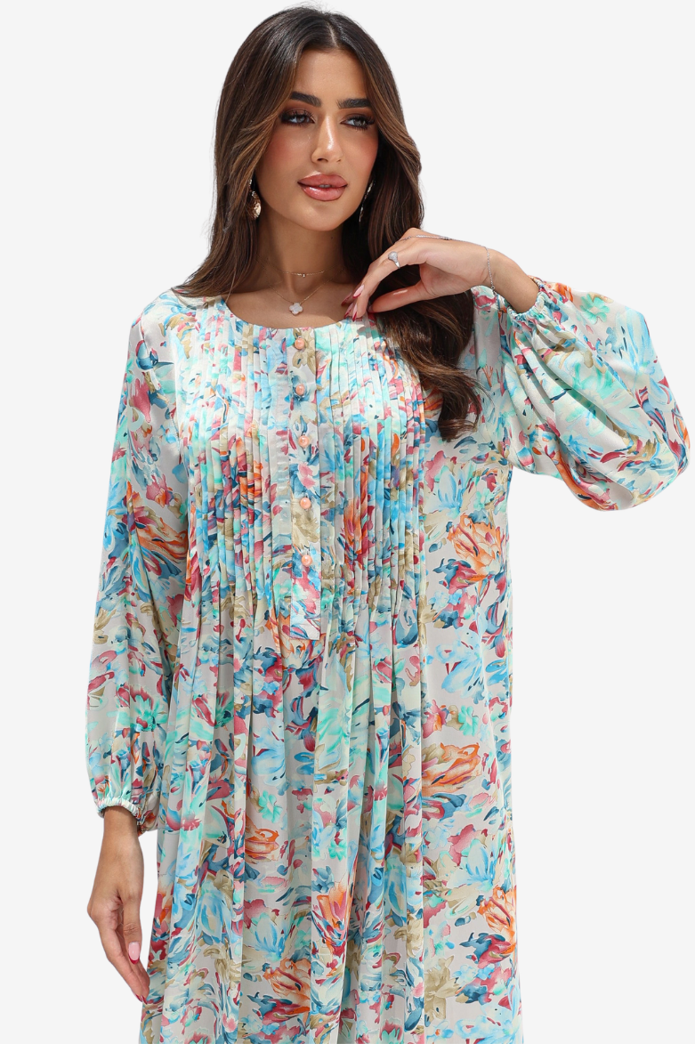 A woman wearing a long-sleeve, pleated tunic dress with a colorful abstract floral print on a light beige background, set against a plain light gray studio backdrop.