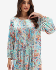 A woman wearing a long-sleeve, pleated tunic dress with a colorful abstract floral print on a light beige background, set against a plain light gray studio backdrop.
