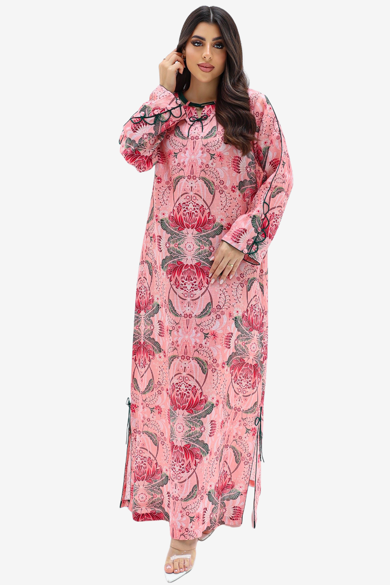 A woman modeling a long-sleeved pink maxi dress with a red and green floral pattern, lace-up side details, and a round neckline, set against a plain light gray background.