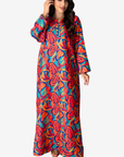 A woman modeling a long-sleeved maxi dress with a vibrant, multicolored abstract pattern in red, orange, blue, and yellow on a light gray background.