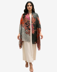 A woman modeling a lightweight open-front shawl with a complex paisley and floral print in orange, red, pink, and gray, worn over a cream-colored kurta and trousers.