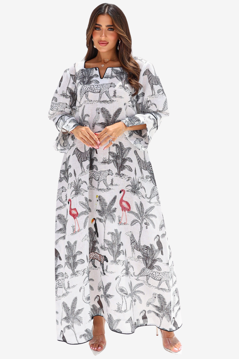 A woman modeling a long white maxi dress with a black and gray animal and palm tree print, featuring pink flamingo accents, set against a plain light gray background.