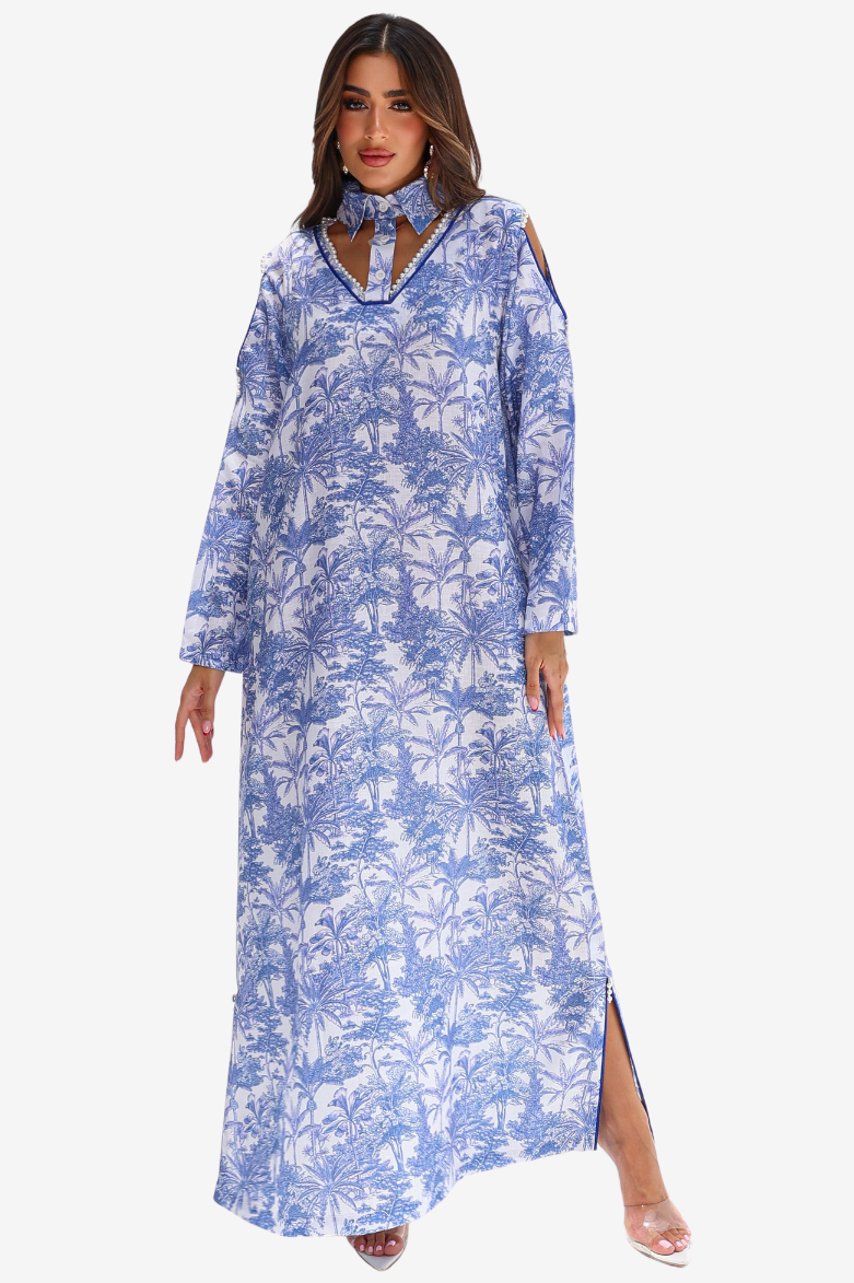 A woman modeling a long-sleeve blue and white tropical palm tree print maxi dress with a high neck, cutout details at the chest, and side slits, set against a plain light gray background.
