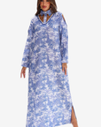 A woman modeling a long-sleeve blue and white tropical palm tree print maxi dress with a high neck, cutout details at the chest, and side slits, set against a plain light gray background.