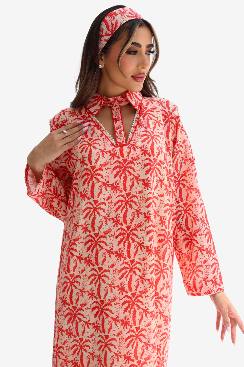 A woman wearing a long-sleeved red and cream palm tree print dress with a keyhole tie-neck, matching headband, and earrings, set against a plain light gray background.