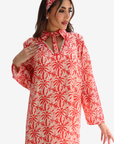 A woman wearing a long-sleeved red and cream palm tree print dress with a keyhole tie-neck, matching headband, and earrings, set against a plain light gray background.