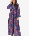 A woman modeling a long-sleeved, floor-length floral dress with a purple, blue, and pink pattern against a plain light gray background.
