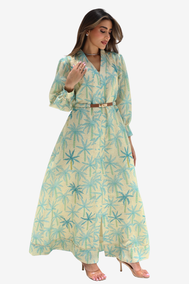 A woman modeling a long-sleeve maxi dress with a light yellow base and a blue palm tree pattern, featuring a matching belt at the waist.