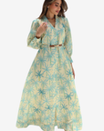 A woman modeling a long-sleeve maxi dress with a light yellow base and a blue palm tree pattern, featuring a matching belt at the waist.