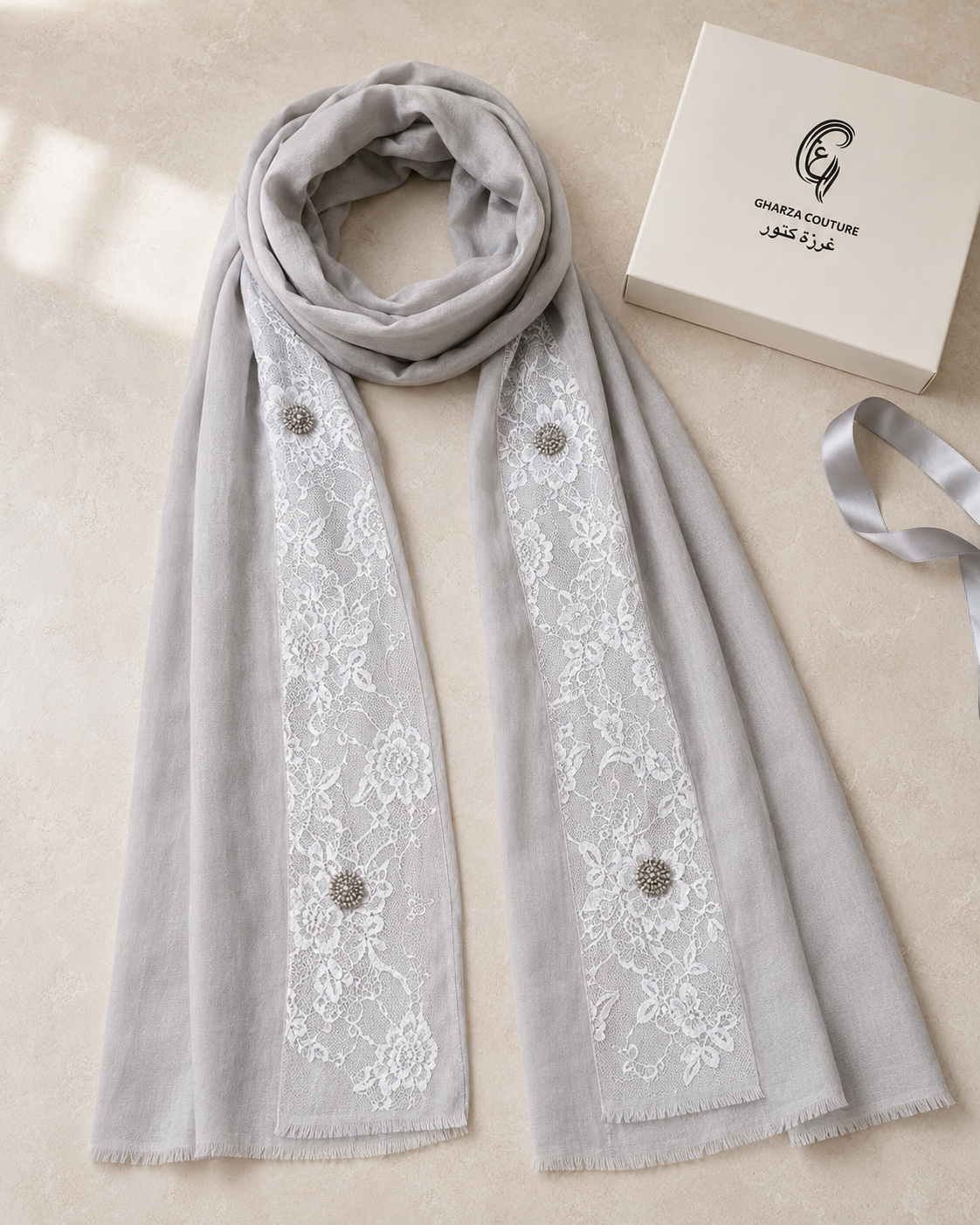A light gray scarf with white floral lace panels and decorative metallic buttons, displayed on a beige surface next to a white box labeled Chanel Couture and a matching gray ribbon.