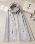 A light gray scarf with white floral lace panels and decorative metallic buttons, displayed on a beige surface next to a white box labeled Chanel Couture and a matching gray ribbon.