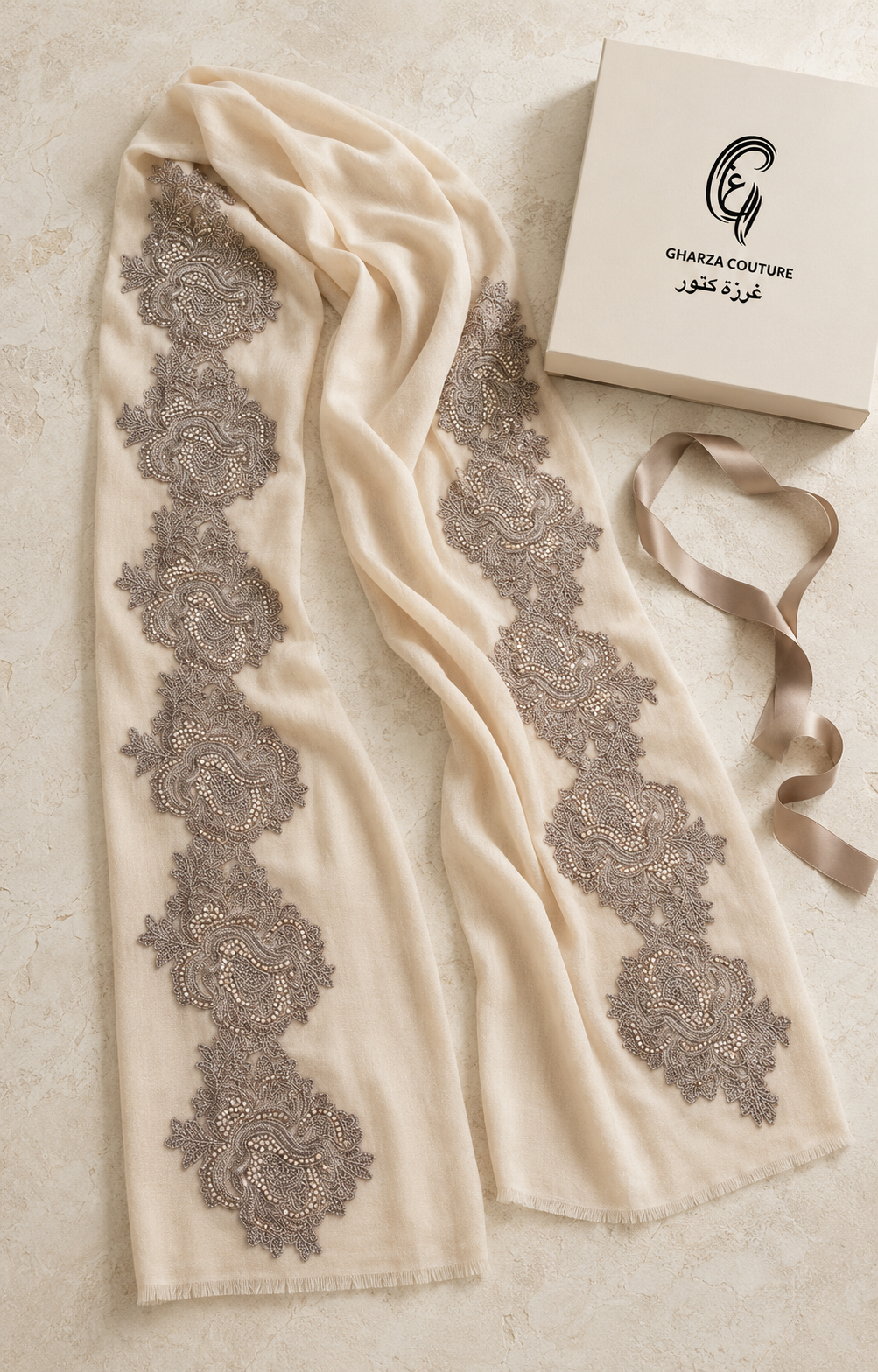 An off-white scarf with vertical gray floral lace patterns, displayed next to its cream-colored Ghada Couture box and a matching ribbon on a textured beige surface.