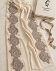 An off-white scarf with vertical gray floral lace patterns, displayed next to its cream-colored Ghada Couture box and a matching ribbon on a textured beige surface.