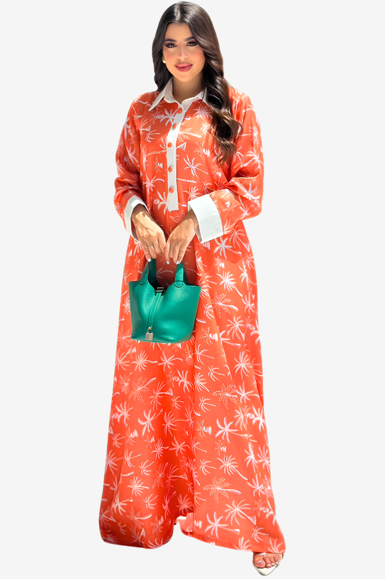 A woman modeling a long-sleeved orange maxi dress with a white palm tree pattern, featuring a white collar and cuffs, paired with a small green handbag against a light gray background.