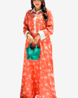 A woman modeling a long-sleeved orange maxi dress with a white palm tree pattern, featuring a white collar and cuffs, paired with a small green handbag against a light gray background.