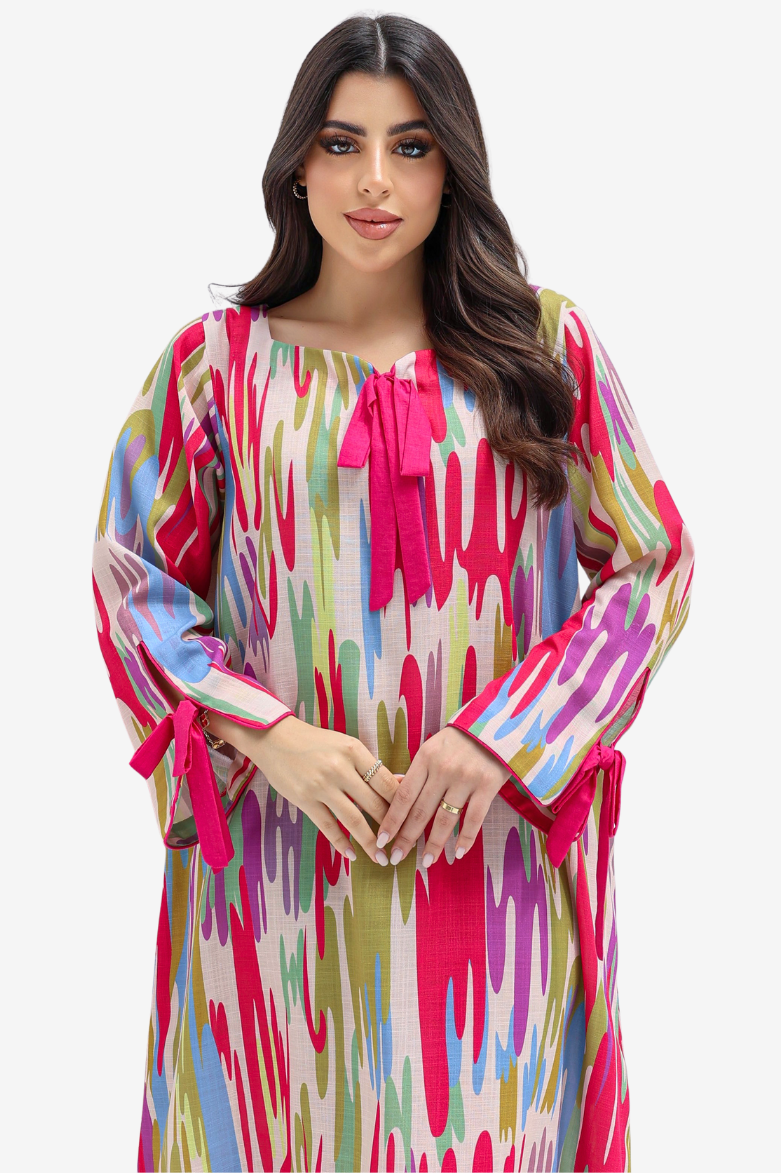 A woman modeling a long-sleeved, loose-fitting tunic dress with a vibrant multicolor abstract print. The dress features a bright pink tie-neck bow and matching pink tie details on the cuffs, set against a plain light gray studio background.