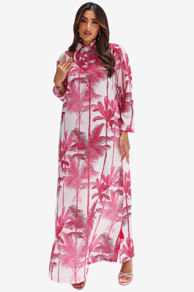 A woman modeling a long-sleeved, high-neck maxi dress with a pink and red palm tree print on a white background, set against a plain light gray studio backdrop.