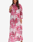 A woman modeling a long-sleeved, high-neck maxi dress with a pink and red palm tree print on a white background, set against a plain light gray studio backdrop.
