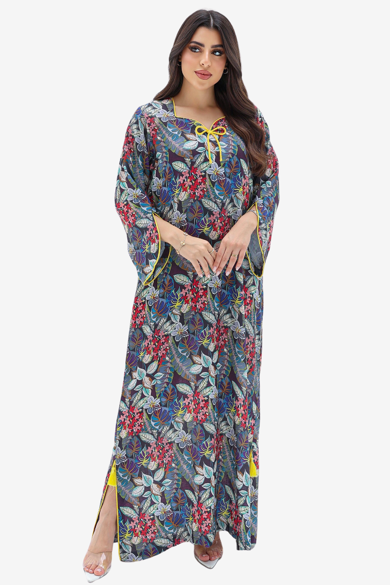 A woman modeling a long, loose maxi dress with a dense floral and botanical print. The dress features a muted blue-gray base color with prominent red, pink, and green floral motifs, accented by yellow tie details at the neckline and yellow side slits.