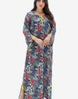 A woman modeling a long, loose maxi dress with a dense floral and botanical print. The dress features a muted blue-gray base color with prominent red, pink, and green floral motifs, accented by yellow tie details at the neckline and yellow side slits.