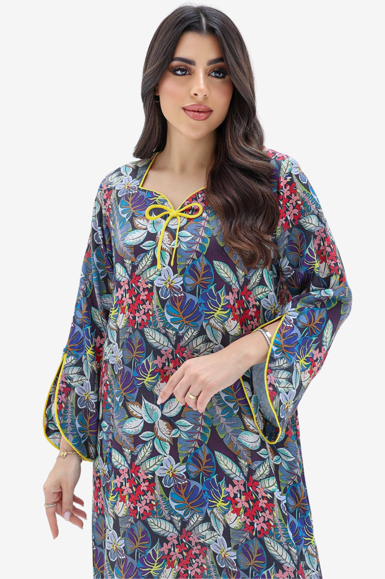 A woman modeling a long-sleeve kaftan dress with a dense floral and botanical print in blue, red, green, and yellow, featuring a yellow tie at the neckline and yellow piping details.