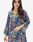 A woman modeling a long-sleeve kaftan dress with a dense floral and botanical print in blue, red, green, and yellow, featuring a yellow tie at the neckline and yellow piping details.