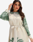 A woman modeling a cream-colored dress with green tropical leaf prints on the sleeves, collar, and hem, featuring a matching fabric tie belt at the waist.