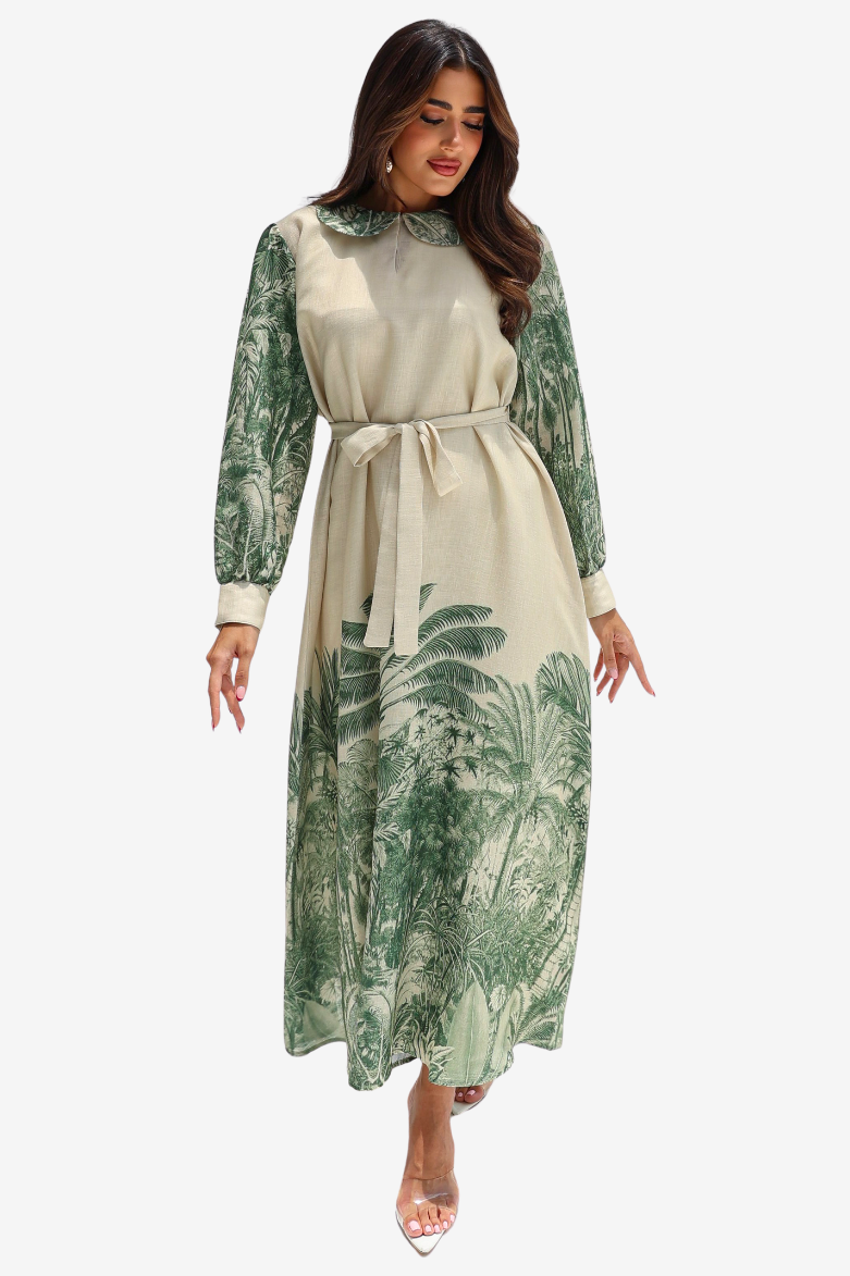 A woman modeling a long-sleeve midi dress with a cream base and green tropical leaf print, featuring a matching fabric tie belt at the waist, set against a plain light gray background.